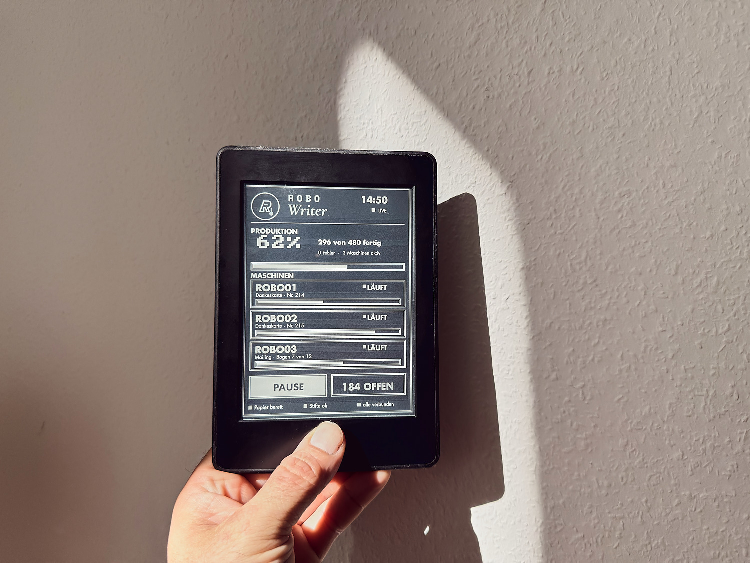 Hand hält E-Ink-Display mit Produktionsstatus: drei Schreibroboter aktiv, 62 Prozent fertig, 296 von 480 Aufträgen, Maschinenraum Stuttgart